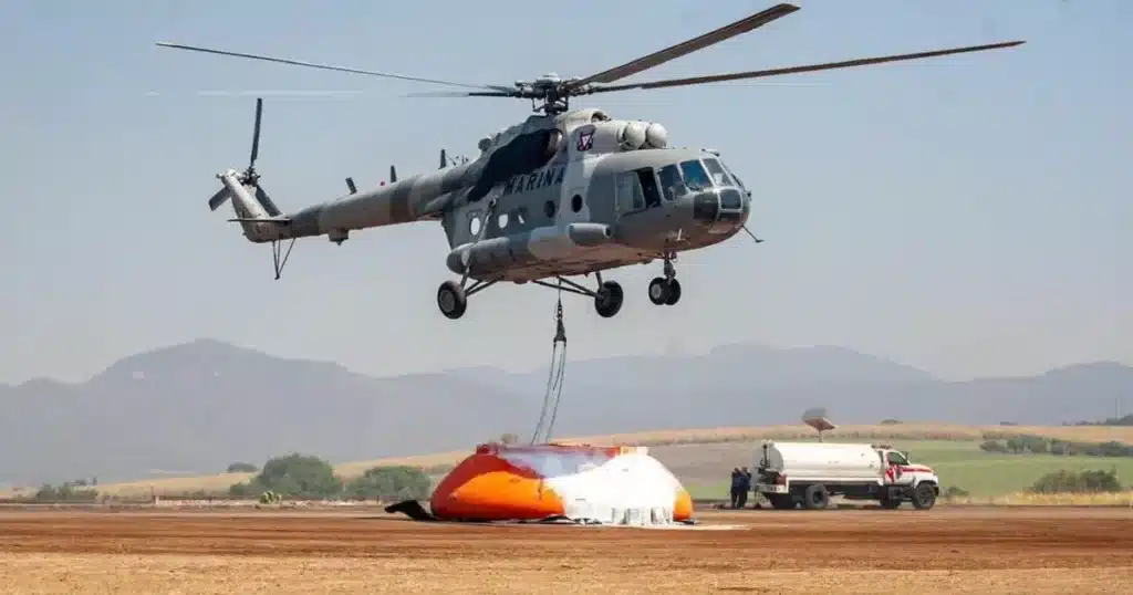 Un helicóptero gris planea sobre el suelo, llevando un gran cubo de agua naranja y blanco. Al fondo, se ven un camión cisterna y montañas bajo un cielo despejado.