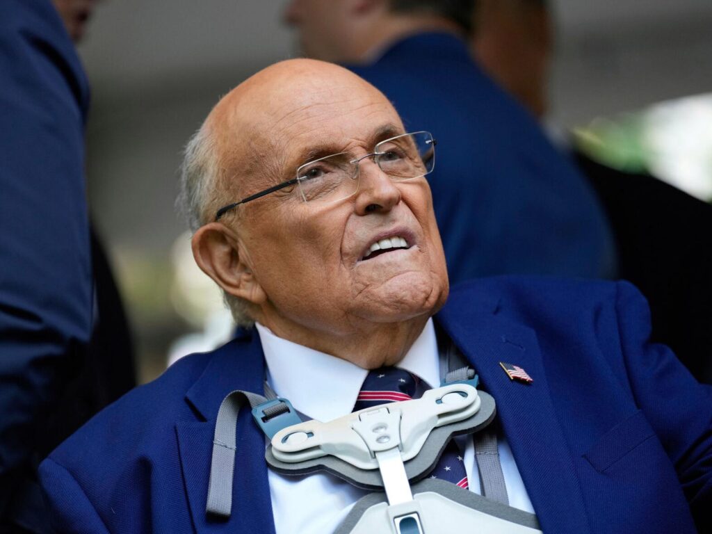 Un hombre mayor con gafas y un aparato ortopédico en la cabeza, vestido con traje azul, camisa blanca y corbata a rayas, mira hacia arriba con expresión neutra. En el fondo se ven borrosas personas con trajes oscuros.