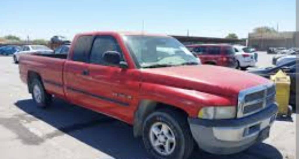 Una camioneta roja con cabina extendida está aparcada en un solar en un día soleado, rodeada de otros vehículos. La camioneta muestra algunos signos de desgaste y tiene llantas plateadas y molduras negras.