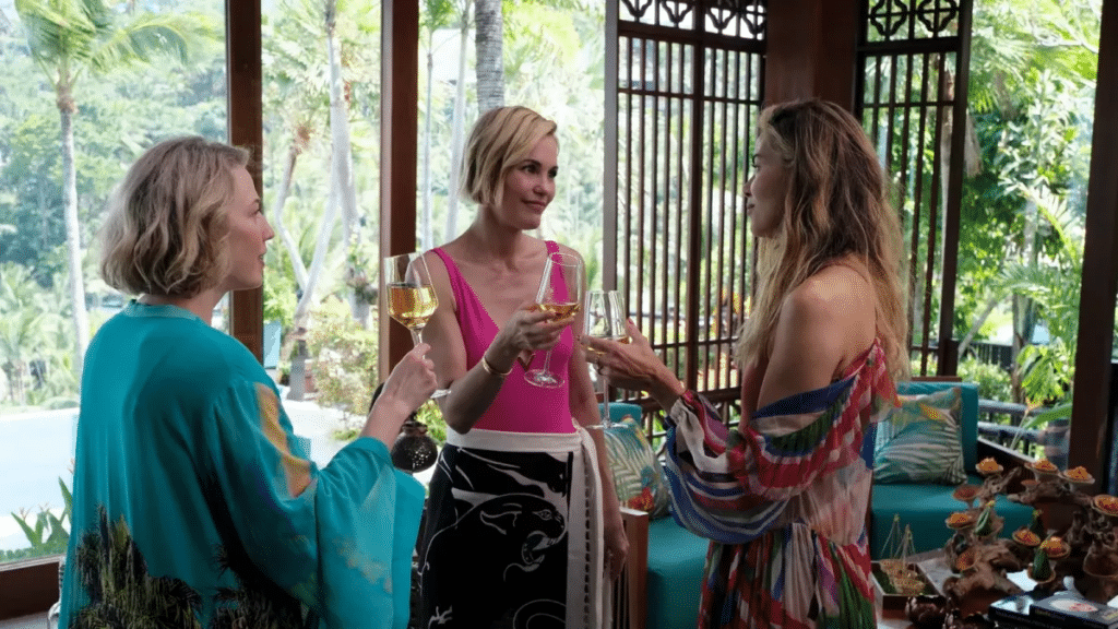 Tres mujeres permanecen juntas en el interior, junto a grandes ventanales, sosteniendo vasos de vino blanco y conversando. Llevan ropa colorida y veraniega, y a través de las ventanas se ve vegetación en el exterior.