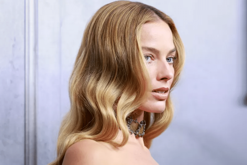 Una mujer de pelo rubio largo y ondulado y piel clara mira hacia un lado. Lleva un collar gargantilla con joyas y maquillaje natural, de pie sobre un fondo borroso de color claro.